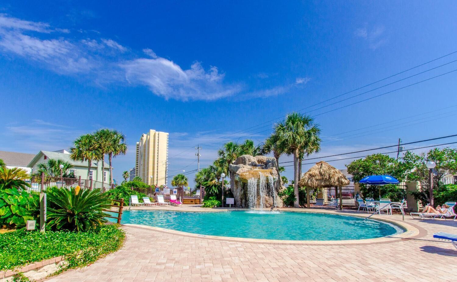 A pool area with a waterfall feature, lounge chairs, palm trees, and a thatched-roof structure.