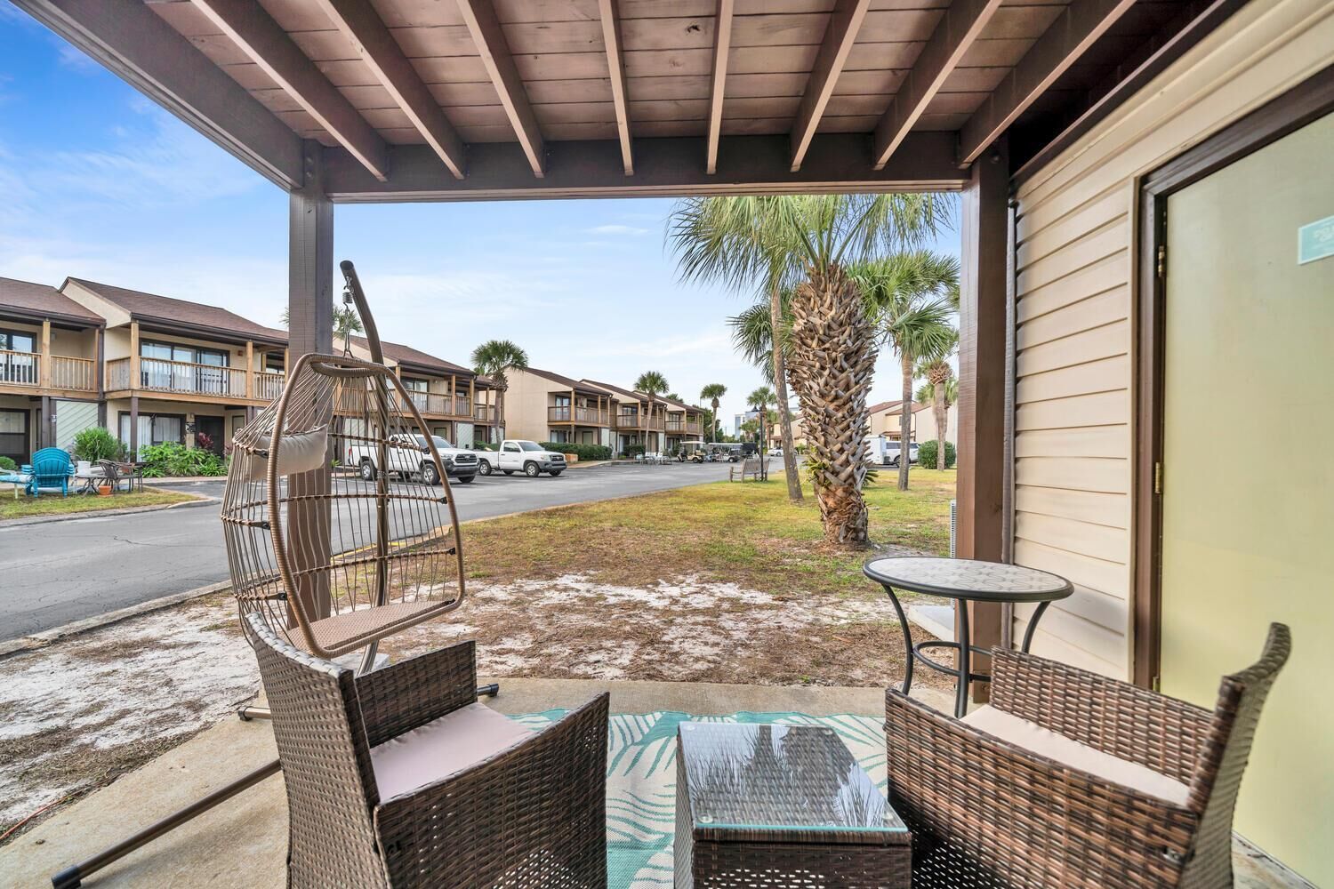 A covered outdoor seating area with wicker furniture, a view of a street with palm trees, and a building with balconies in the background.