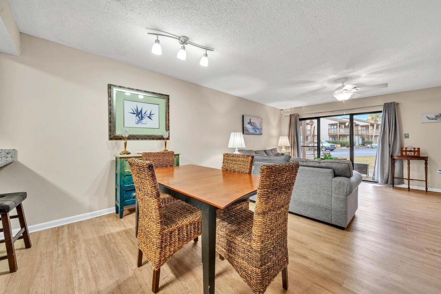 A dining area with a wooden table and wicker chairs, a grey sofa, and a view of the outside through a glass door.