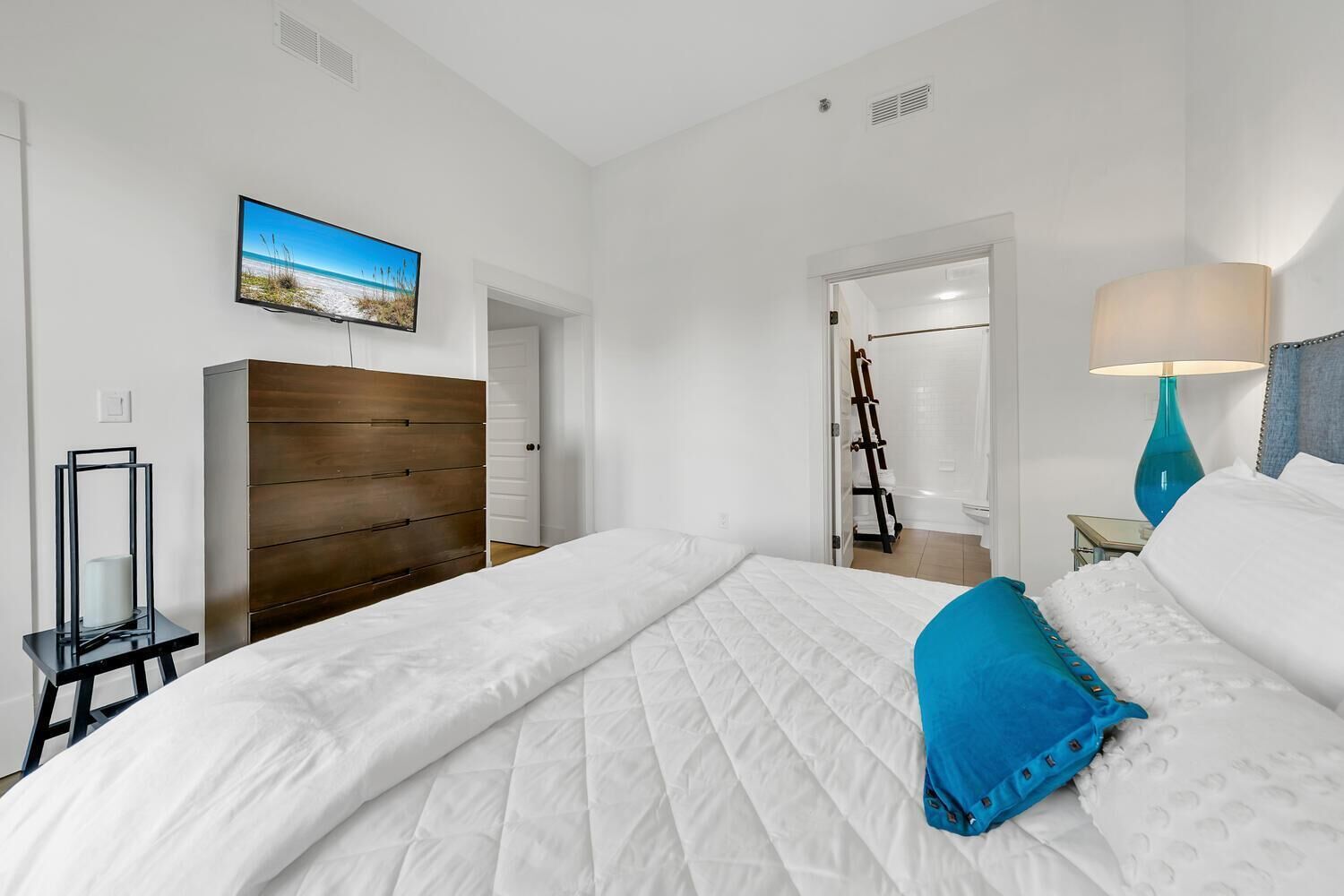 A hotel room with a large bed, a wooden headboard, a TV on the wall, a bathroom visible through an open door, and a nightstand with a lamp.