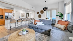 A modern living room with a grey sofa, a coffee table, and a kitchen area with wooden cabinets and a refrigerator. - Redfish Village M1222 (Santa Rosa Beach)