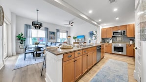 A modern kitchen with wooden cabinets, a central island, stainless steel appliances, and a dining area with a table and chairs. - Redfish Village M1222 (Santa Rosa Beach)