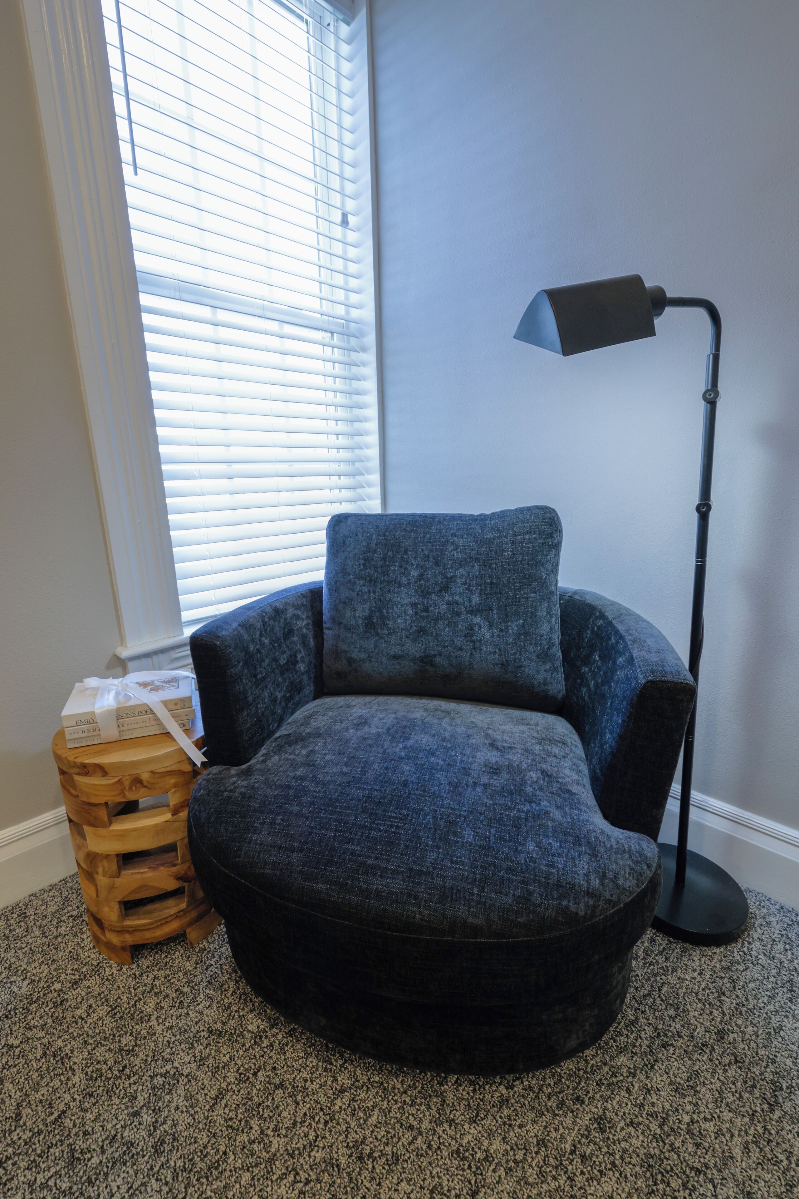 Une pièce d’angle avec un fauteuil bleu, une lampe sur pied, une caisse en bois et une fenêtre avec des stores.