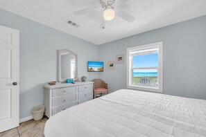 Slaapkamer met een groot bed, een dressoir, een stoel, een plafondventilator, een raam met uitzicht op het strand en een deur.