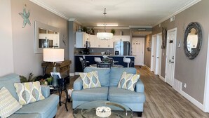 A living room with a blue sofa, a glass coffee table, and a kitchen in the background. - Ocean Villa 1303 (Panama City Beach)