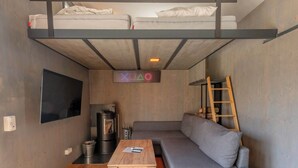 Un espacio habitable compacto con una cama litera, un sofá, un televisor, una mesita de centro y una escalera de madera.