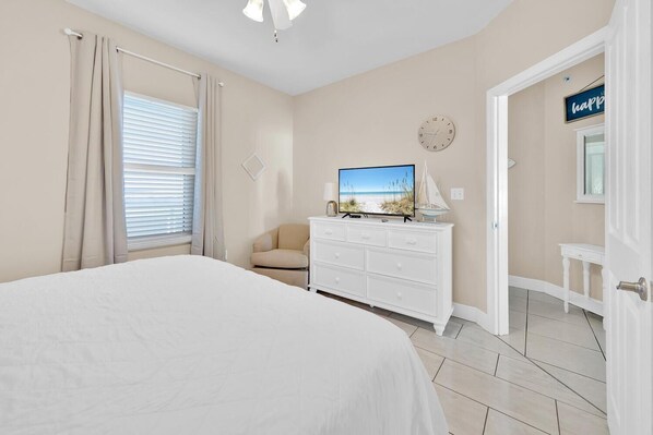 A bedroom with a bed, a TV on a dresser, a chair, a clock, and a door with a 'happy' sign. - Celadon 1103 - Beach Paradise (Panama City Beach)