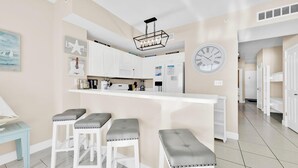 A modern kitchen with white cabinets, a central island with bar stools, and a clock on the wall. - Celadon 1103 - Beach Paradise (Panama City Beach)