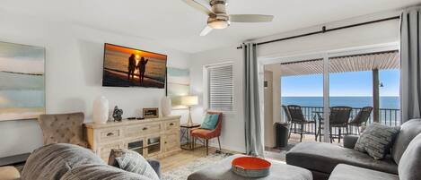 A living room with a large window overlooking the sea, a ceiling fan, and a view of the ocean from the balcony.