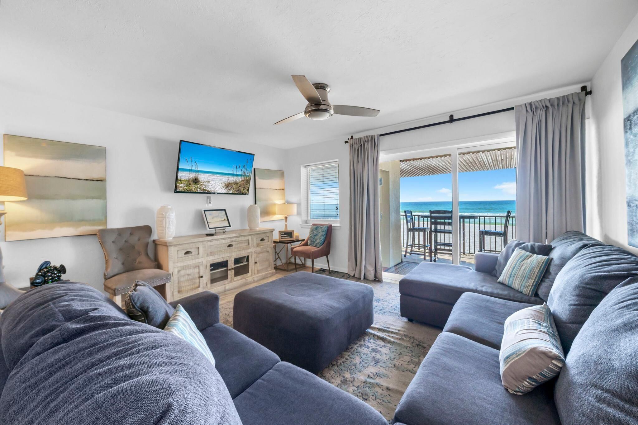 A modern living room with a large gray sectional sofa, a small wooden cabinet, a television, and a view of the ocean through large windows.