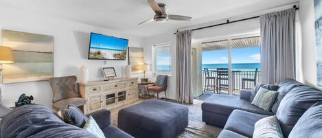 A modern living room with a large gray sectional sofa, a small wooden cabinet, a television, and a view of the ocean through large windows.