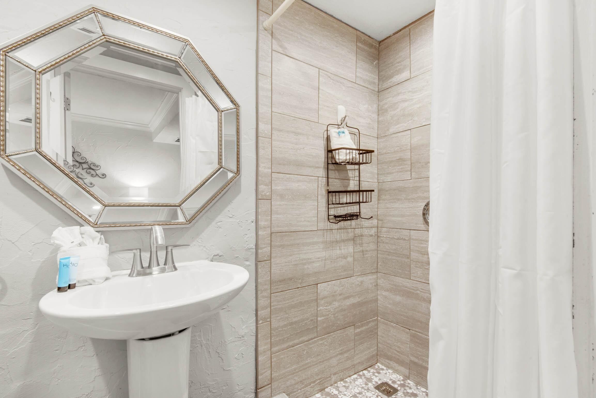 Un baño con un lavabo blanco, un espejo grande con marco dorado y una zona de ducha con azulejos color beige.