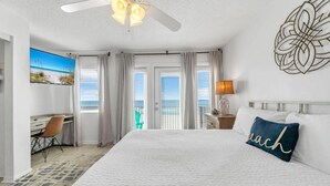 A hotel room with a large bed, a desk, a chair, and a view of the ocean through the windows. - The Sand Trap- Costa del Sol C-9 (Miramar Beach)