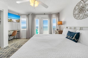 A hotel room with a large bed, a desk, a chair, and a view of the ocean through the windows. - The Sand Trap- Costa del Sol C-9 (Miramar Beach)