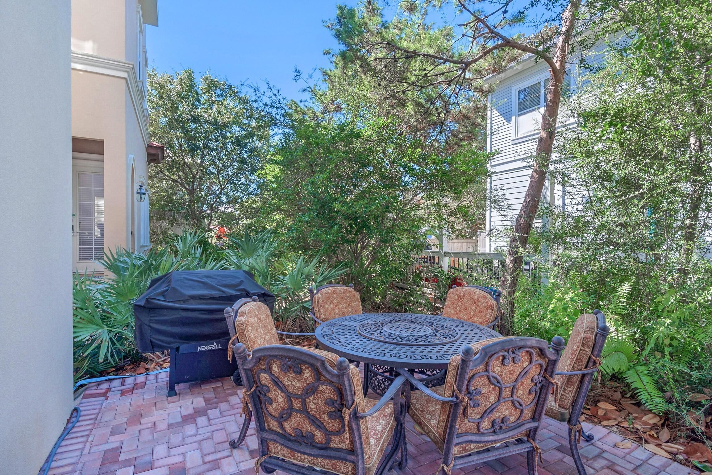 An outdoor patio with a round metal table and patterned chairs, surrounded by lush greenery and a covered grill.