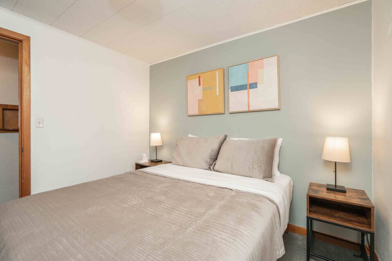 Un lit bien fait, recouvert d’une couverture beige, deux tables de chevet avec des lampes et deux tableaux abstraits accrochés au mur.