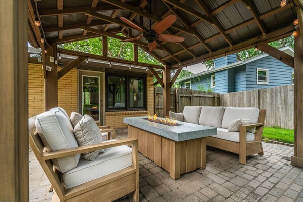 Eine überdachte Terrasse mit einem Holztisch und zwei Sofas, einem Deckenventilator und Blick auf ein Haus im Hintergrund.