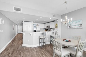 A modern kitchen with white cabinetry, a central island with stools, and a dining area with a round table and chairs. - Majestic Beach Towers 2-803 (Panama City Beach)