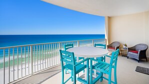 A balcony with a white table and turquoise chairs, overlooking a beach with a clear blue sky and ocean.