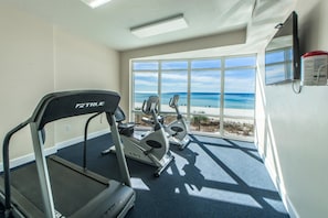 Un gimnasio con cinta de correr, elíptica y bicicleta estática, ventanas amplias con vista a la playa y una lámpara en el techo.