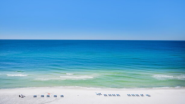 A beach with white sand and clear blue water. There are several blue lounge chairs and a red umbrella on the sand.