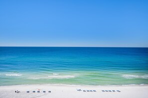 Ein Strand mit weißem Sand und klarem, blauem Wasser. Mehrere blaue Liegestühle und ein roter Sonnenschirm liegen im Sand.