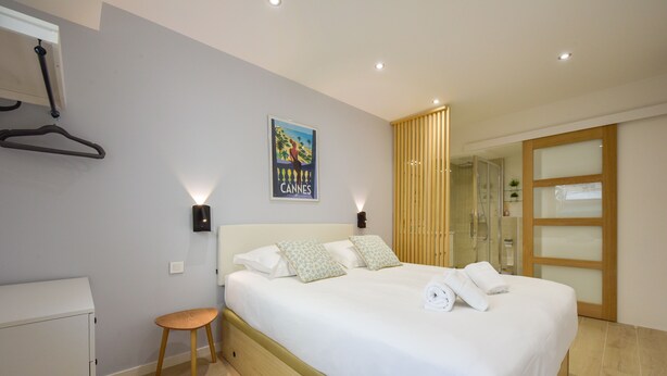 A neatly arranged bedroom with a bed, bedside tables, a small stool, a wardrobe, and a poster on the wall. - Upscale 3-room apartment, 300m from the beaches (Cannes)