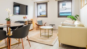 A modern living room with a sofa, a dining table with chairs, a TV mounted on the wall, and large windows with curtains. - Upscale 3-room apartment, 300m from the beaches (Cannes)