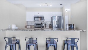 A modern kitchen with white cabinets, stainless steel appliances, granite countertops, and three bar stools. - Pointe of View 407- Penn's Paradise (Miramar Beach)