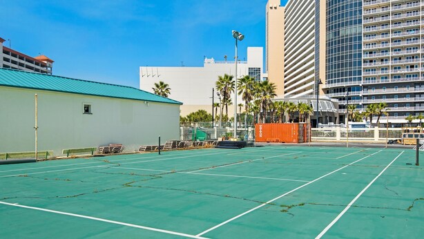 An outdoor tennis court with a green surface, surrounded by buildings and palm trees. - Sunbird 810e (Panama City Beach)