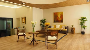 Een moderne woonkamer met een houten plafond, een gele bank, een bijzettafeltje en twee stoelen. Aan de muur hangt een schilderij en rechts staat een potplant.