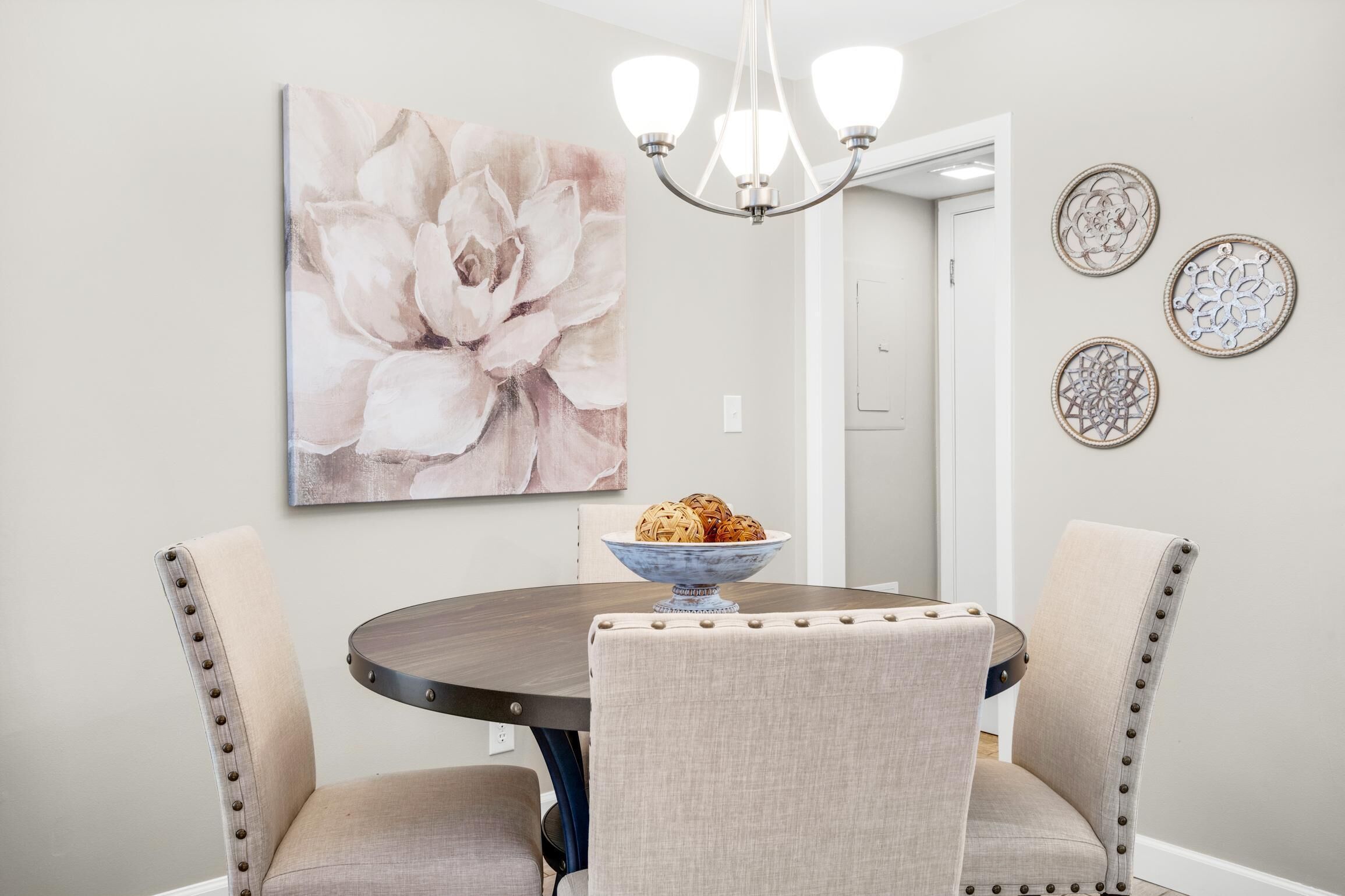 Un comedor con una mesa redonda, cuatro sillas tapizadas, una araña y un cuadro grande con motivos florales en la pared.