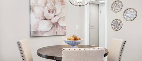 Un comedor con una mesa redonda, cuatro sillas tapizadas, una araña y un cuadro grande con motivos florales en la pared.
