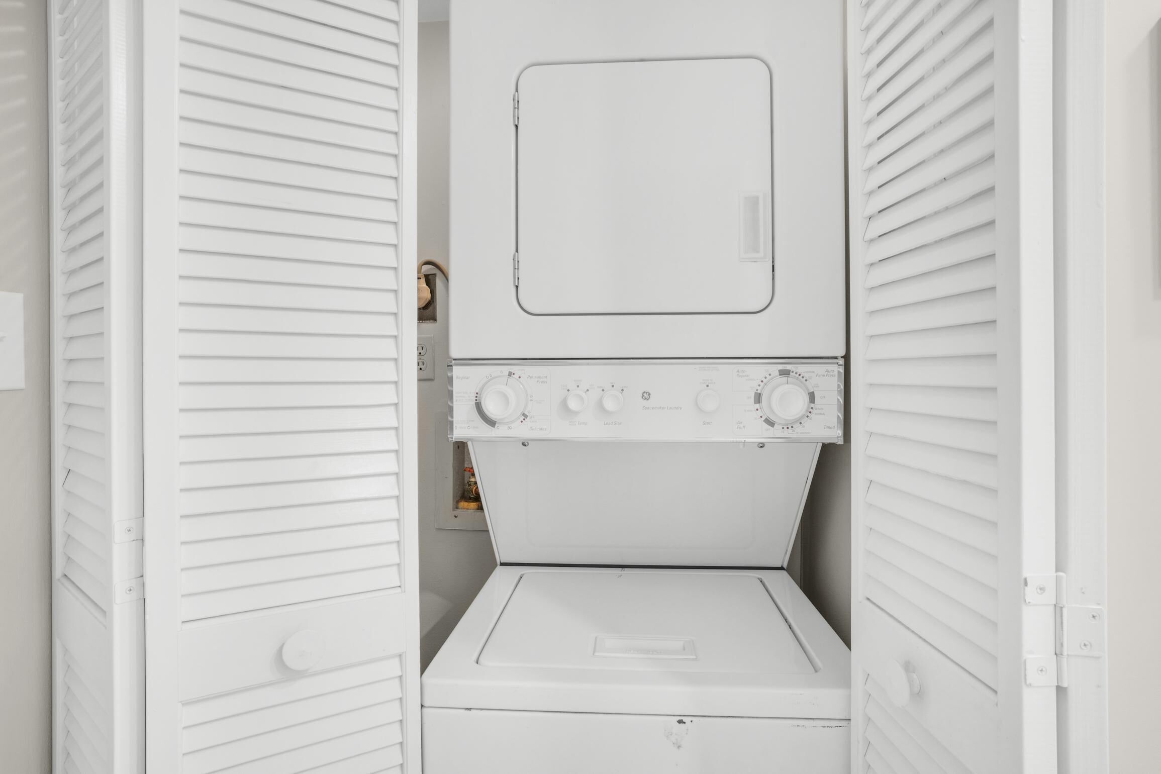 Une buanderie blanche équipée d’un lave-linge et d’un sèche-linge à chargement frontal, d’un panneau de commande avec des boutons et d’une petite étagère sur laquelle se trouve une bouteille.