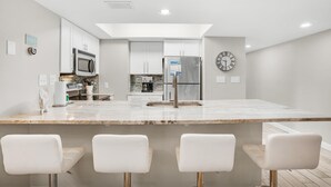 A modern kitchen with a central island, white bar stools, and stainless steel appliances. - Hidden Dunes Beach Cottage 208- Dune Our Thing (Miramar Beach)