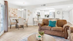 A modern living room with a brown leather sofa, a dining area with a round table, and a kitchen in the background. - Hidden Dunes Beach Cottage 208- Dune Our Thing (Miramar Beach)