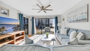 A living room with a large TV, a ceiling fan, a sofa, and a coffee table. - Edgewater Golf Villa 3308 (Panama City Beach)