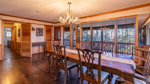 A spacious dining room with a large wooden table and ornate chairs, a chandelier, and large windows offering a view of the outdoors.