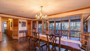 A spacious dining room with a large wooden table and ornate chairs, a chandelier, and large windows offering a view of the outdoors.