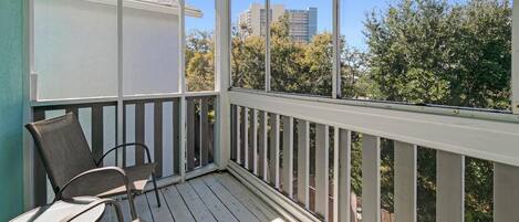Un balcon extérieur couvert, équipé de deux chaises et d’une table, avec des balustrades en bois et une vue sur les arbres et les bâtiments.
