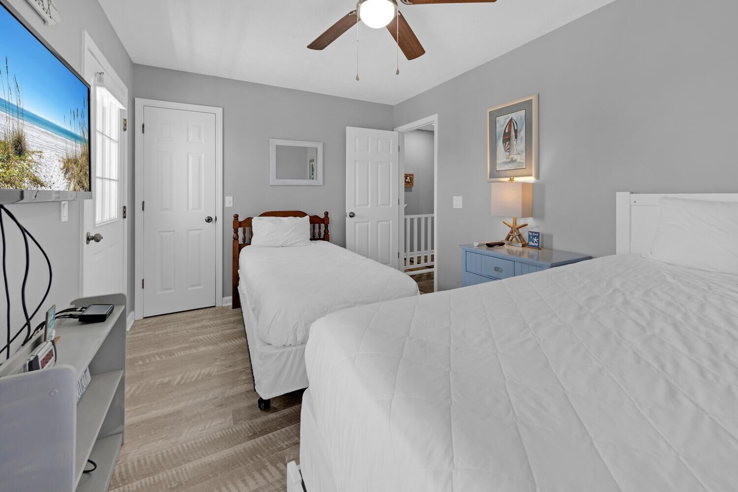 Une chambre avec deux lits, un ventilateur de plafond, un bureau avec un ordinateur et un téléviseur diffusant une scène de plage.