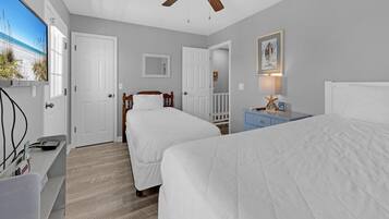 Une chambre avec deux lits, un ventilateur de plafond, un bureau avec un ordinateur et un téléviseur diffusant une scène de plage.