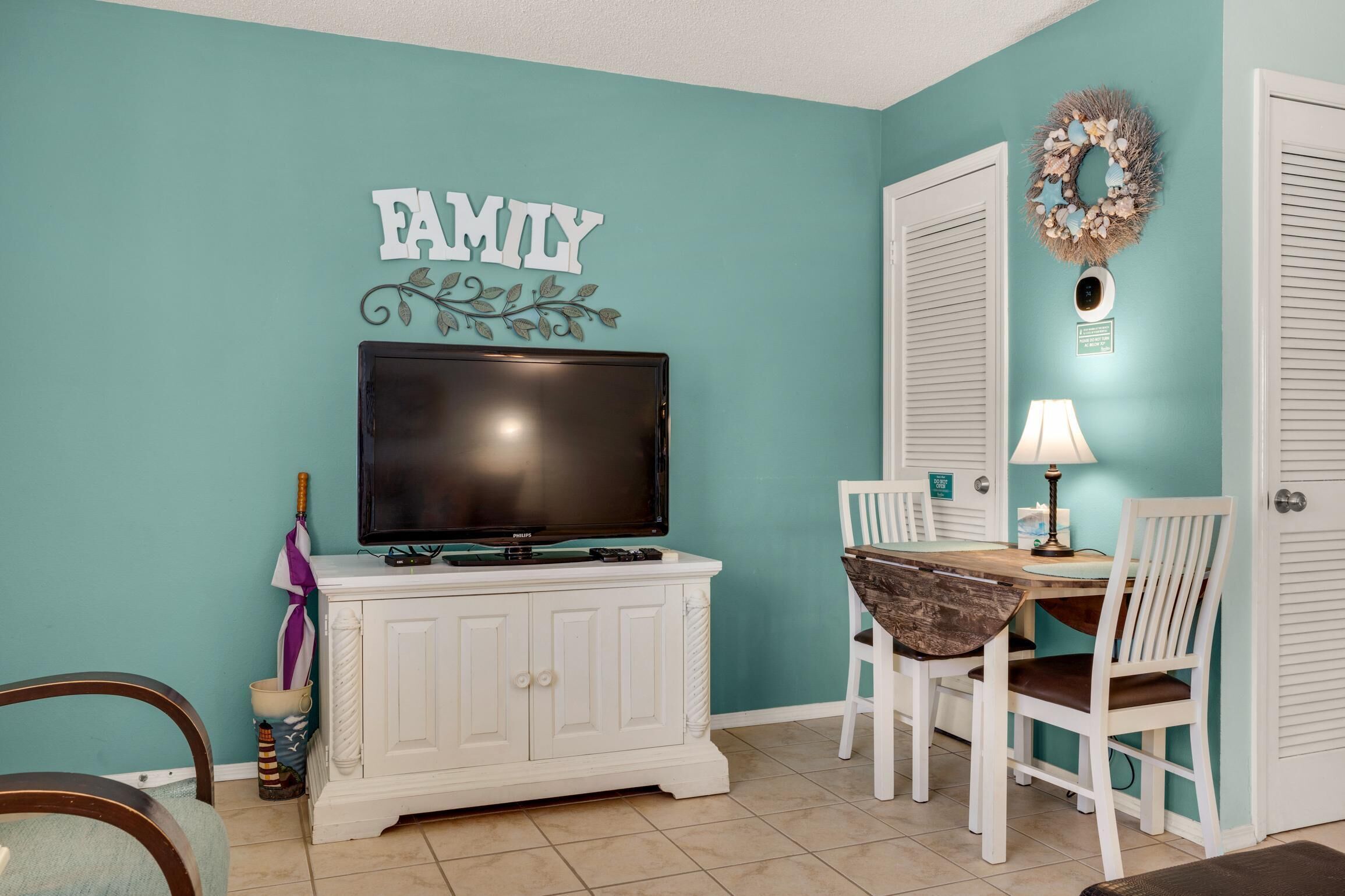 A teal-walled room with a television on a white cabinet, a dining table with chairs, and a wreath on the wall.