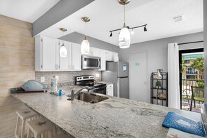 A modern kitchen with white cabinets, a granite countertop, stainless steel appliances, and pendant lighting. - Sandprints A6- Life of Riley (Miramar Beach)