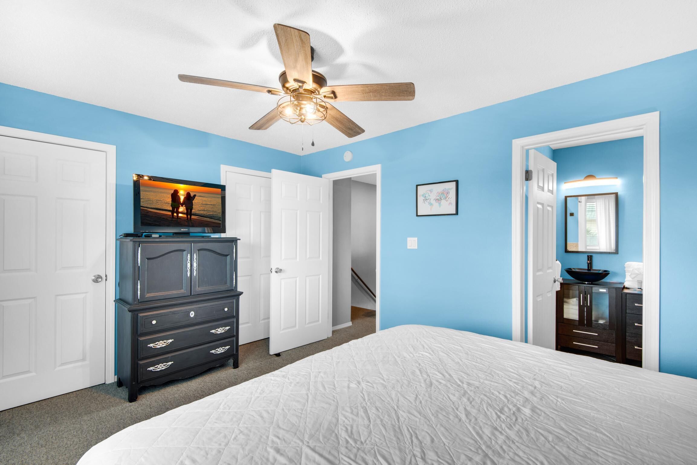 A bedroom with a bed, a dresser, a ceiling fan, a TV, and a bathroom with a sink and mirror.