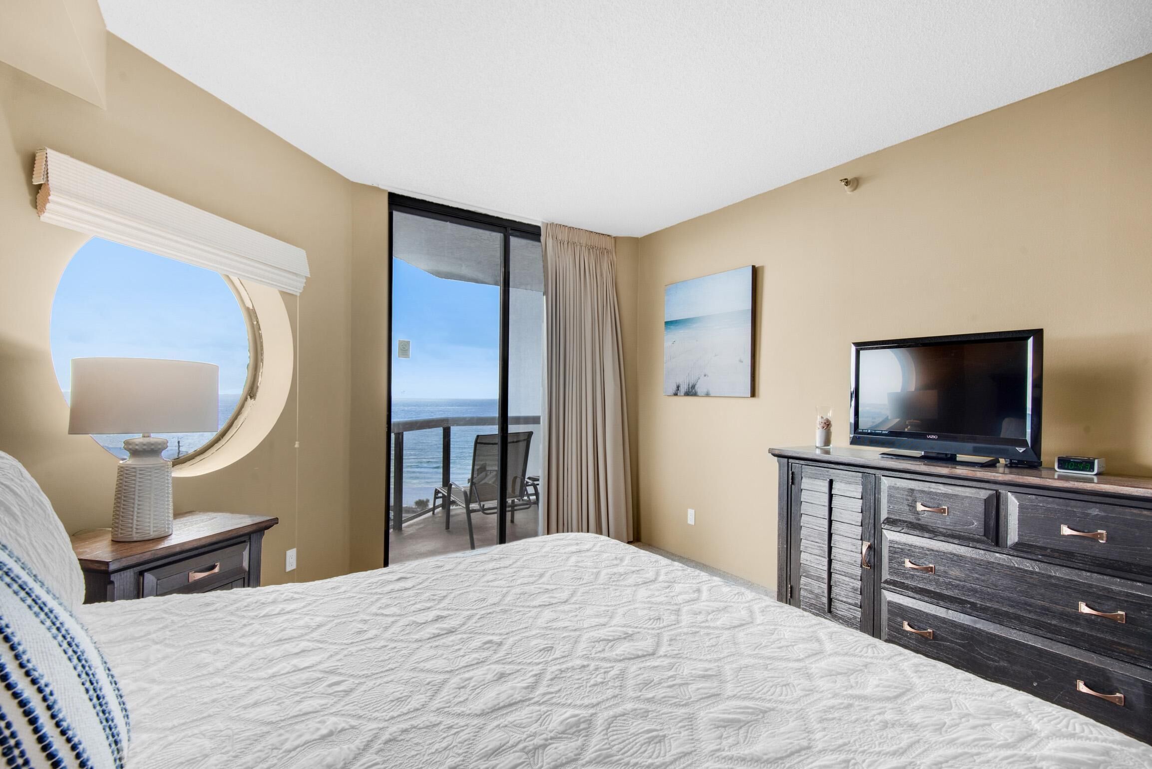 A hotel room with a bed, a TV, a dresser, a lamp, and a view of the ocean.