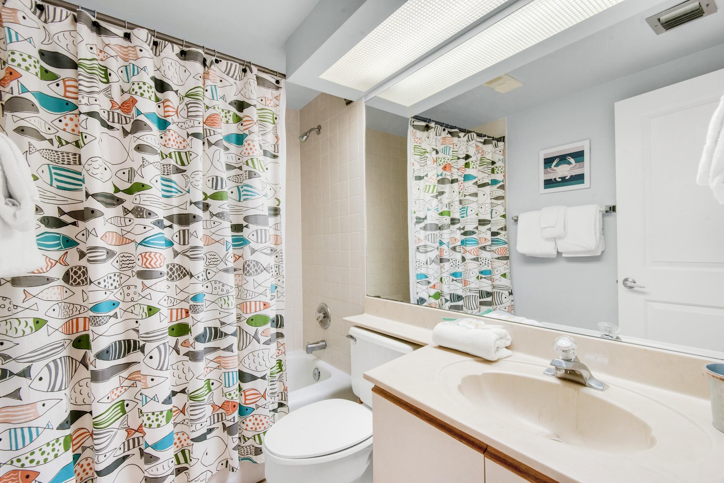 A bathroom with a shower curtain featuring a fish pattern, a white toilet, a sink with a mirror, and a towel rack.