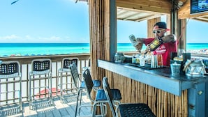 A beachside bar with a view of the ocean, metal chairs, and a bartender preparing drinks.