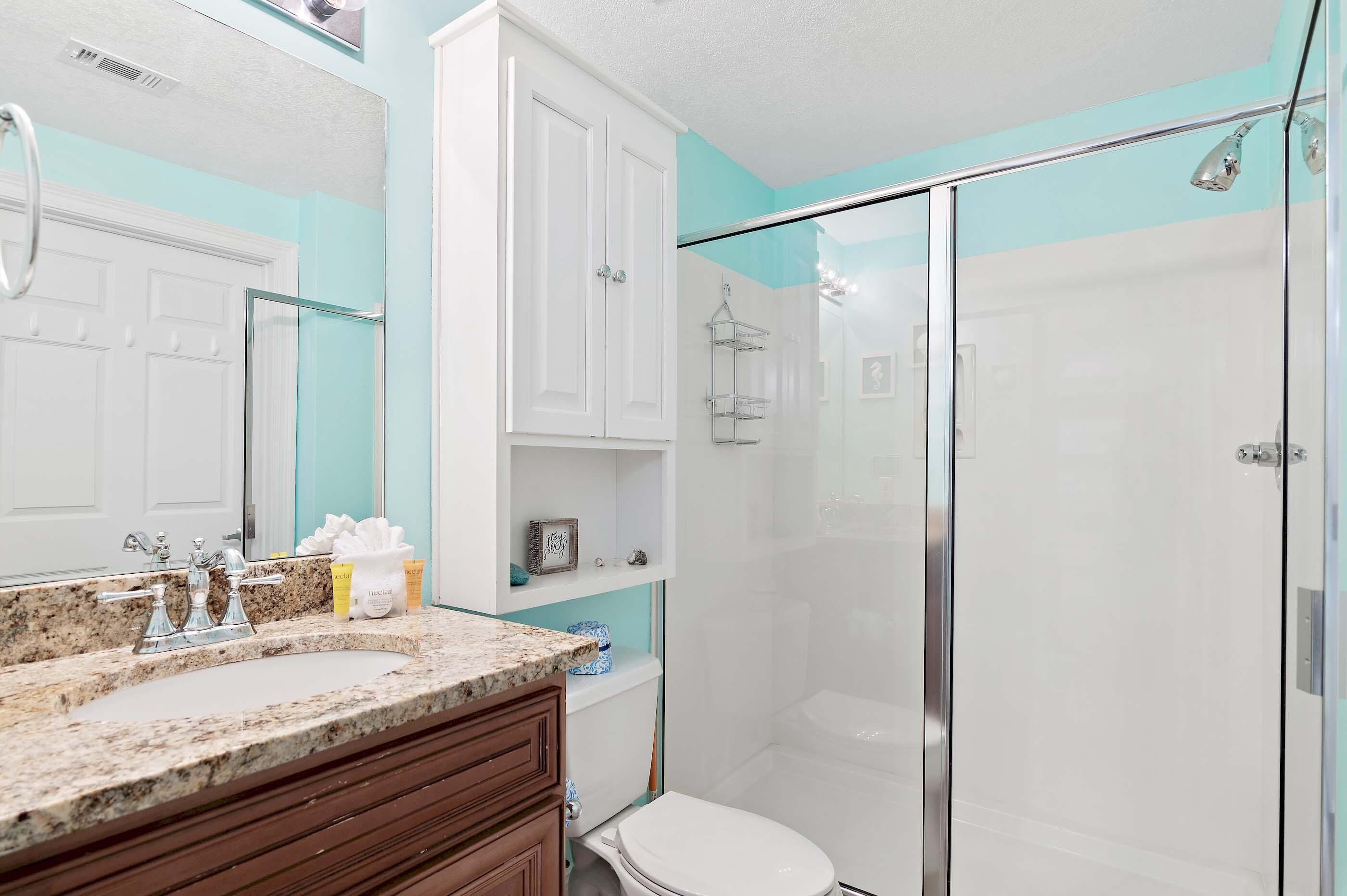 Un baño con encimera de granito, armarios blancos, cabina de ducha de vidrio y un inodoro blanco.