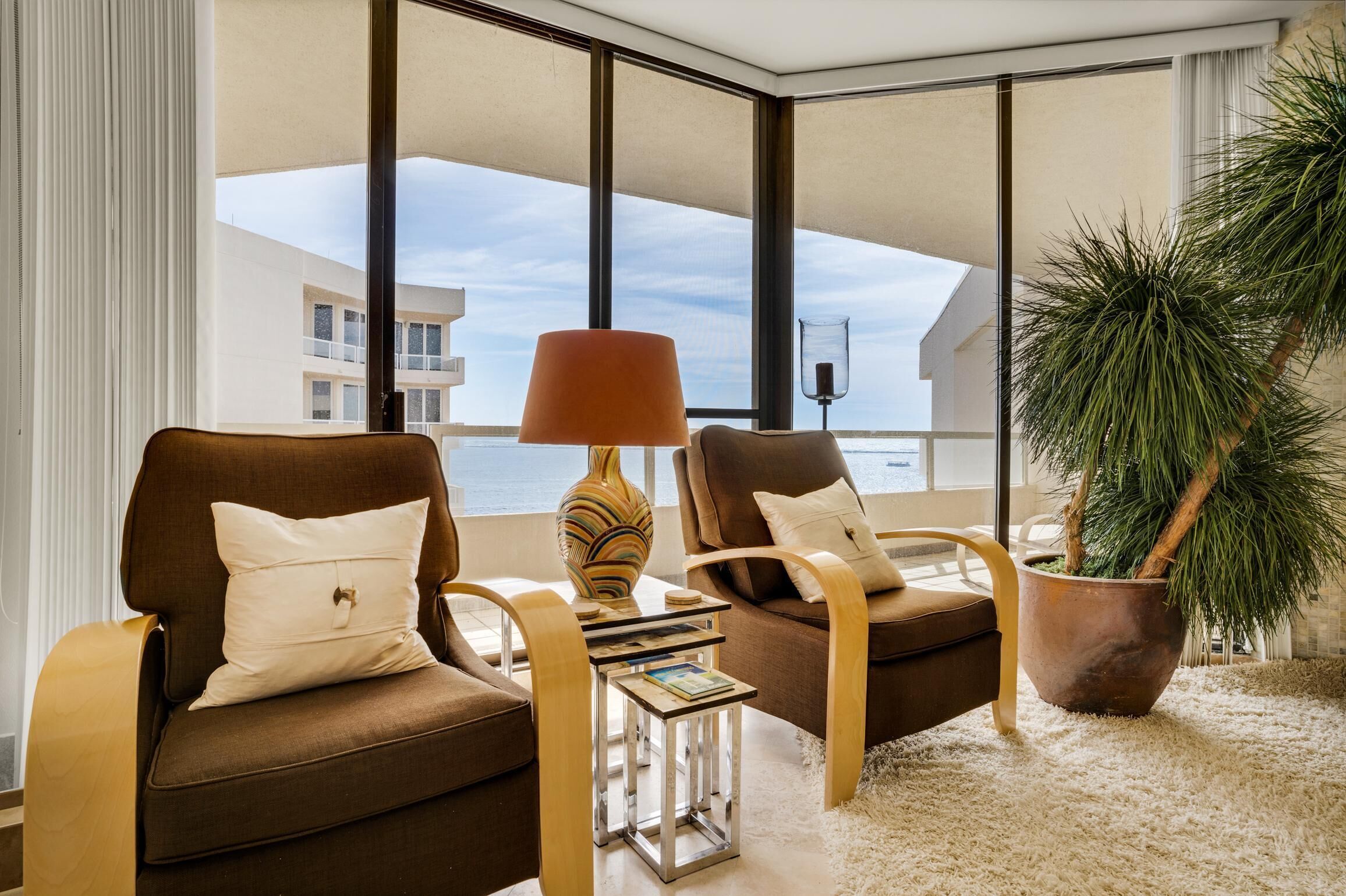A modern living room with two brown armchairs, a side table with a lamp, a potted plant, and a view of a building through large windows.